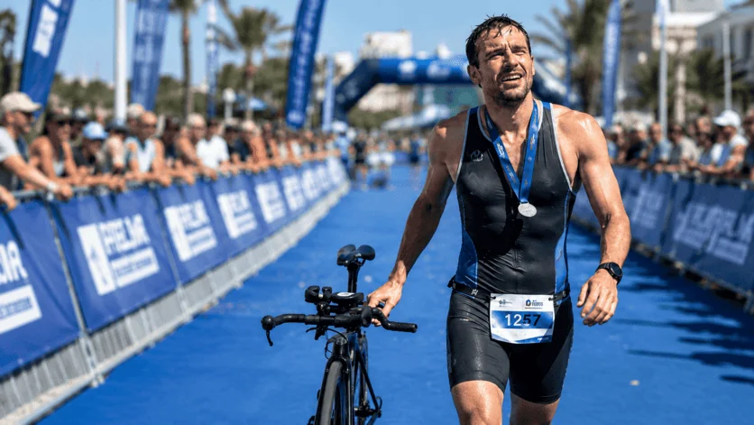 Triatleta cruzando la meta empujando su bicicleta, con la medalla puesta
